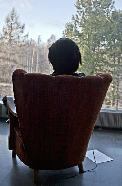 A person sitting in an armchair facing a large window looking at a forest listening to headphones.